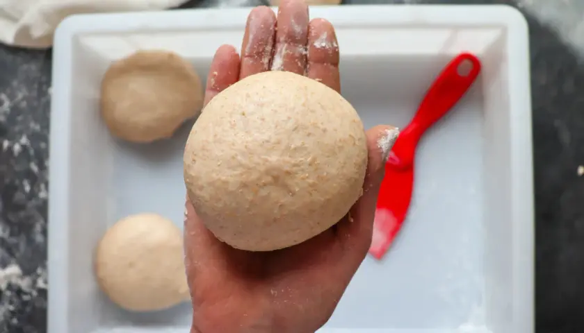 Hand hält klebrigen Pizzateig. Mehr Teiglinge in einer weißen Schale, daneben ein roter Teigschaber.