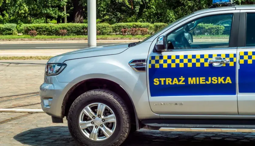 Srebrny samochód Straży Miejskiej z niebiesko-żółtym pasem i napisem "STRAŻ MIEJSKA".