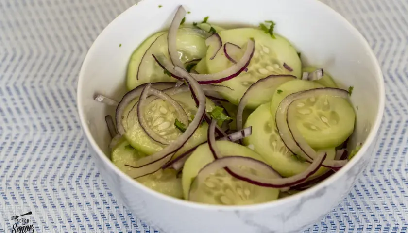 Deliciosa ensalada de pepino con cebolla morada y hierbas frescas, servida en un tazón blanco sobre un mantel azul y blanco.