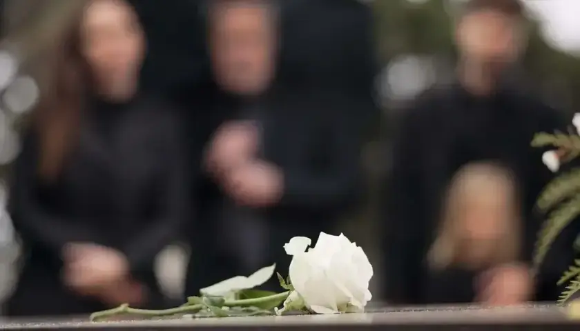Biała róża na kamiennej płycie, w tle zamazane sylwetki żałobników. Czas organizacji pogrzebu jest trudny.