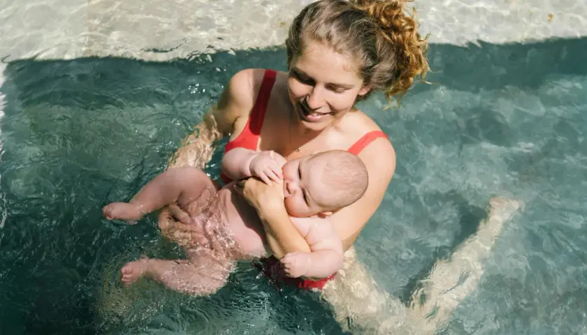 Mamma sorridente tiene in braccio il suo neonato durante un corso di nuoto per neonati in piscina.