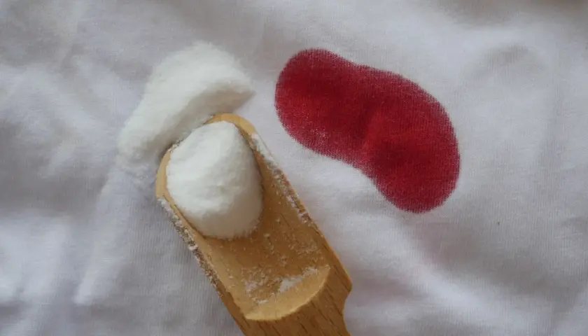 Une cuillère en bois contient une poudre blanche à côté d'une tache rouge sur un tissu blanc. Une tache de sang qui ne part pas au lavage, c'est le cauchemar de tout parent.