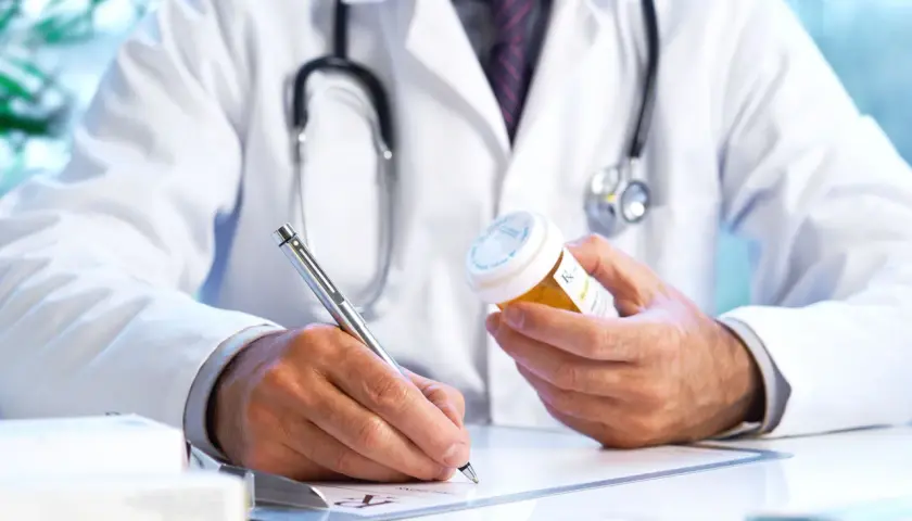 Medico con camice bianco e stetoscopio, scrive su ricetta medica, tenendo in mano un flacone di pillole. Un piano terapeutico medico di base in corso.