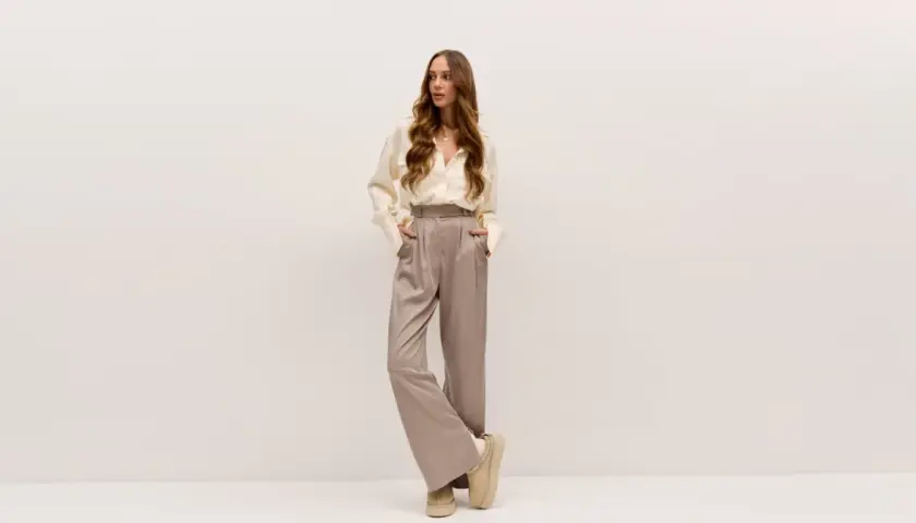 Elegancka kobieta w satynowych spodniach i kremowej koszuli. Idealne buty do teatru? Te wygodne, beżowe botki na platformie dodadzą szyku.