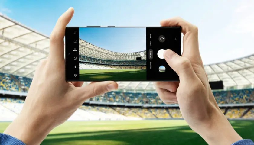 Dwie dłonie trzymają smartfon Samsung S23, robiąc zdjęcie pustego stadionu piłkarskiego.