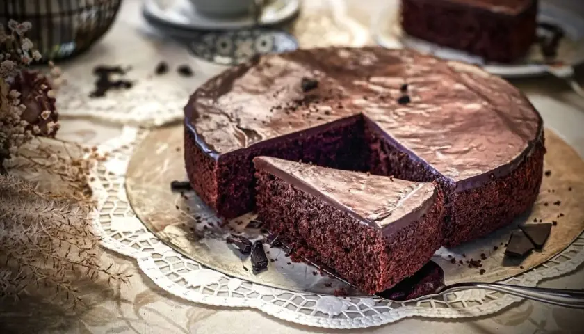 Ein saftiger Schokokuchen mit glänzender Glasur, ein Stück davon auf einem Tortenheber, daneben Schokoladenstücke und eine Kaffeetasse.