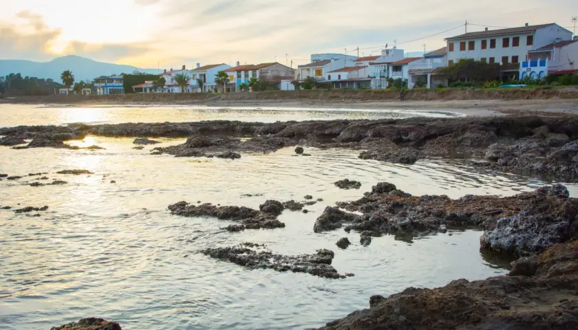 Costa con casas blancas y palmeras, rocas volcánicas en primer plano y el mar reflejando la luz del atardecer. Se divisa una torre la sal a lo lejos.