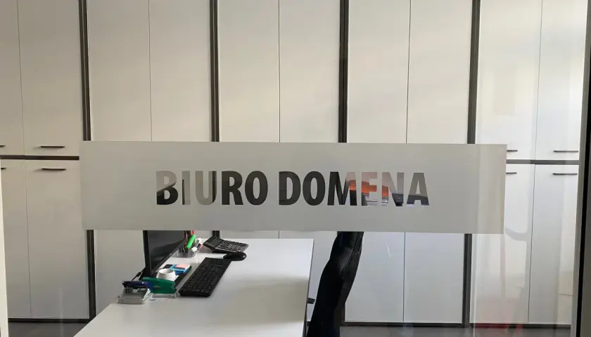 Szklana witryna z napisem "BIURO DOMENA" na tle nowoczesnego biura z białymi szafami.