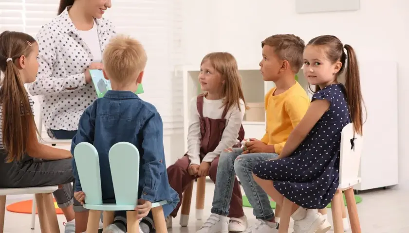 W przedszkolu nauczycielka czyta książkę grupie sześciorga dzieci. Wszyscy siedzą na krzesłach, słuchając uważnie.