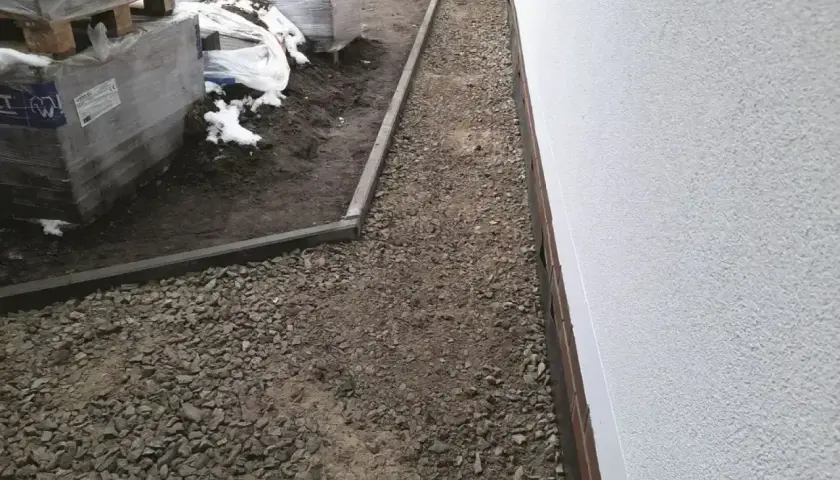 Ułożona opaska przeciwwysadzinowa z kruszywa wokół budynku, obok palety z materiałami budowlanymi.