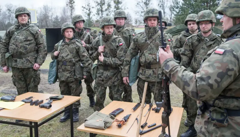 Żołnierze w mundurach ćwiczą, co zabrać na ćwiczenia rezerwy. Na stołach broń, magazynki, nóż i torby.