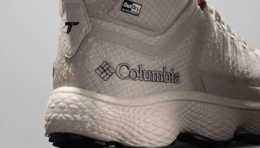 Close-up of a beige Columbia hiking boot with water droplets. The boot features the Columbia logo and "OutDry WATERPROOF" branding, showcasing its durable, water-resistant design.