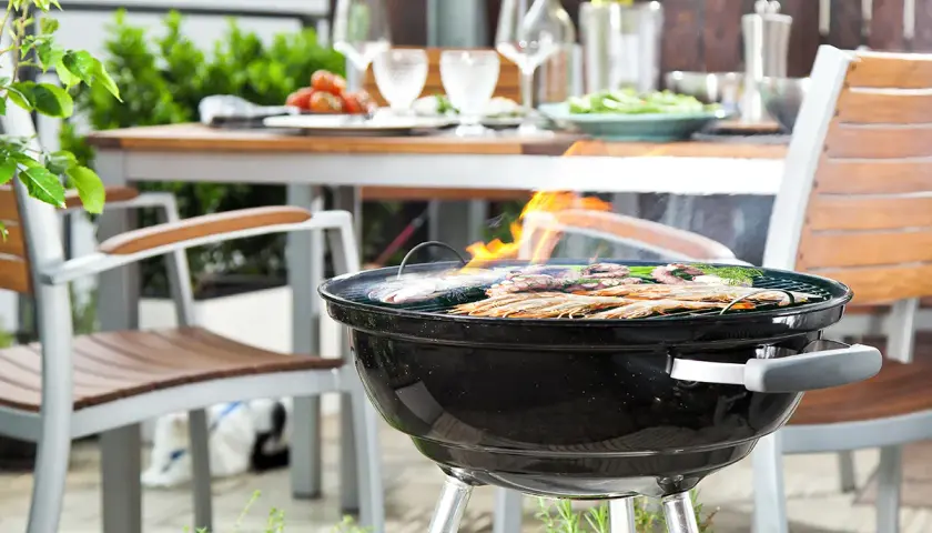 Un barbecue au charbon est allumé, avec des crevettes et du poisson grillant sur la grille. Une table est dressée à l'arrière, prête pour un repas.