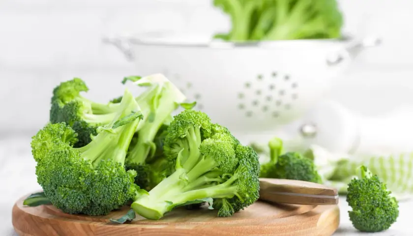 Brocolis frais coupés sur une planche en bois, prêts pour une recette saine. Un égouttoir blanc est visible en arrière-plan.