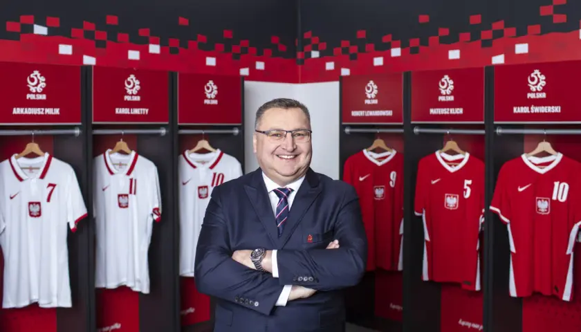 Uśmiechnięty mężczyzna w garniturze stoi przed szatnią reprezentacji Polski. Widać tam koszulki z nazwiskami znanych piłkarzy, tworząc zgrupowanie reprezentacji Polski.