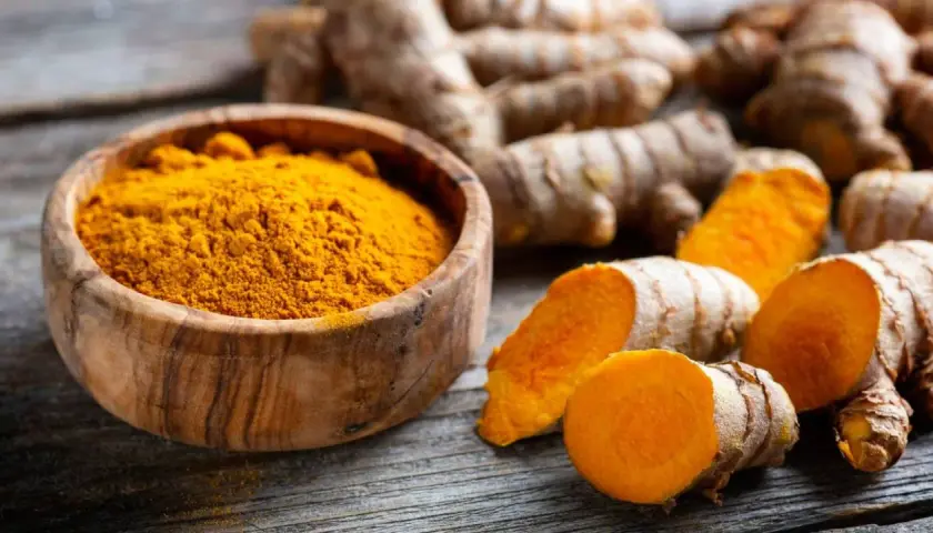 Raíces de cúrcuma y polvo dorado en cuenco de madera, evocando las propiedades de la cúrcuma en cápsulas.