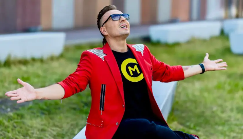 Czadoman w czerwonej marynarce i czarnej koszulce z logo siedzi na ławce z rękami rozłożonymi.