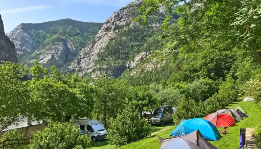 Acampar en este valle es una experiencia de los mejores camping. Tiendas de campaña y furgonetas aparcadas en un prado verde, con imponentes montañas boscosas de fondo.