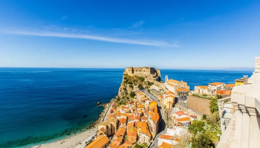 A stunning view of Scilla, Calabria, with its colorful houses cascading down to a beach and the ancient Ruffo Castle perched on a cliff overlooking the turquoise sea. This is what to see in Calabria!