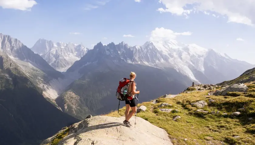 A hiker with a red backpack pauses on a rocky outcrop, enjoying the breathtaking view of the Mont Blanc massif during a challenging hike.