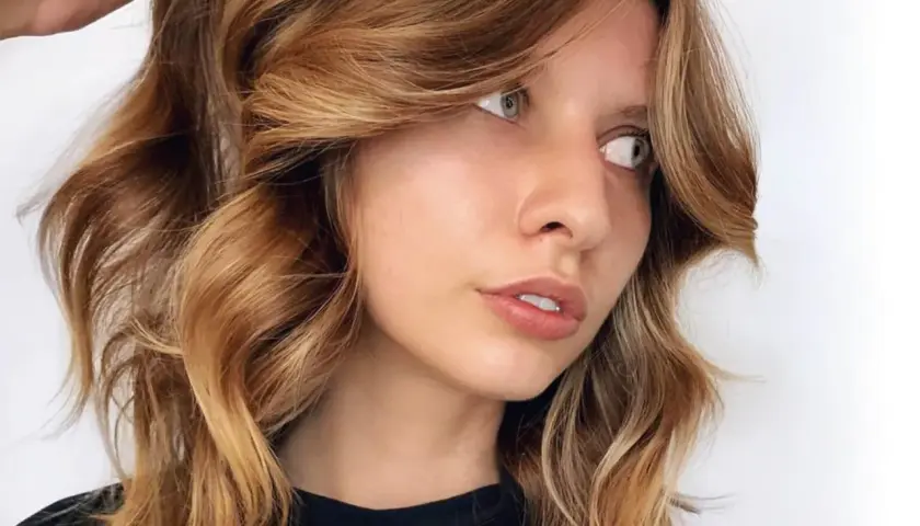 Mujer con cabello castaña iluminada, con ondas suaves y reflejos dorados que resaltan su rostro.