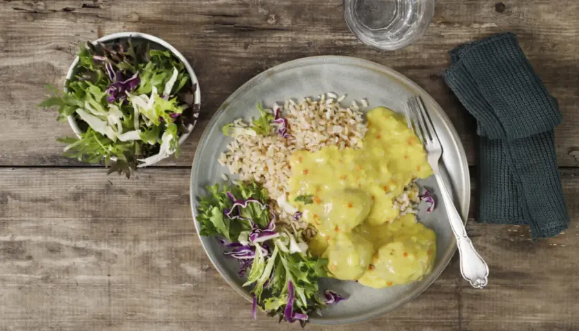 En tallrik med ris, grönsaker och krämiga boller i karry. En skål med sallad står bredvid.