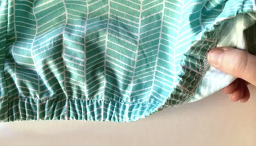 Close-up of a hand demonstrating how to sew in elastic on a teal and white chevron patterned fabric. The elastic creates a gathered, ruffled effect.