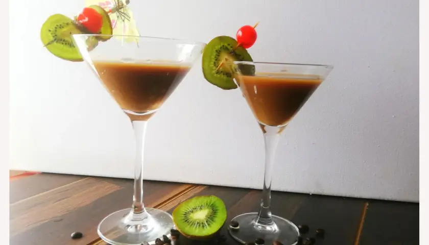 Zwei Martinigläser mit braunem Getränk, garniert mit Kiwi und Kirsche. Kaffeebohnen und eine halbe Kiwi liegen auf dem Tisch. Inspiration für Montenegro Rezepte.
