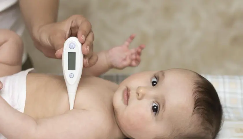 Dłoń rodzica mierzy temperaturę niemowlaka. Maluch leży spokojnie, patrząc w obiektyw.