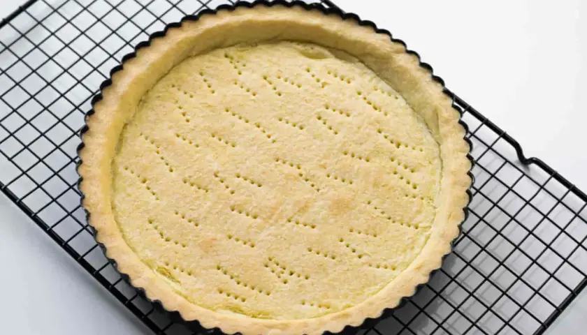 Base di pasta sucrée dorata e forata, pronta per essere farcita, su una griglia nera.