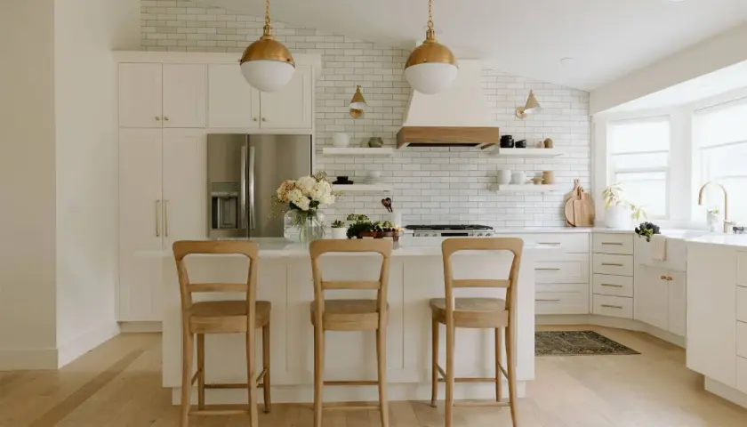 A bright, airy modern farmhouse kitchen with white subway tile, gold pendant lights, and a large island with three wooden stools.