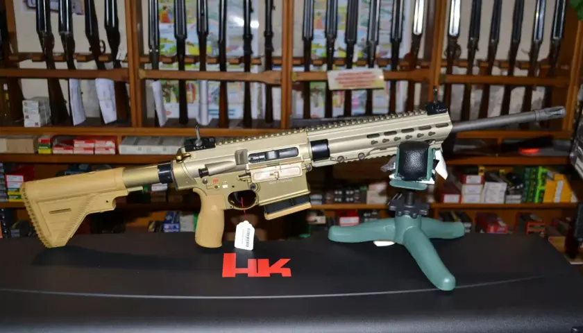 Un fusil HK MR 308, de couleur sable, est présenté sur un support. Derrière, des étagères chargées de fusils et de boîtes de munitions.