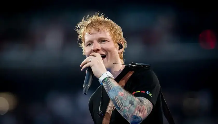 Ed Sheeran śpiewa z mikrofonem, jego ręka ozdobiona kolorowymi tatuażami.