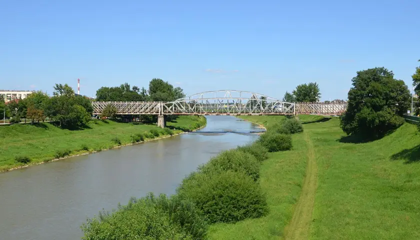 Metalowy most kolejowy nad rzeką w Przemyślu, otoczony zielenią i błękitnym niebem.