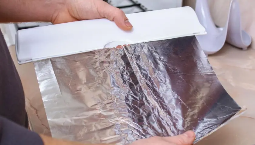 Ręce trzymają biały dozownik, z którego wysuwa się matowa strona folii aluminiowej. Gotowanie w kuchni.