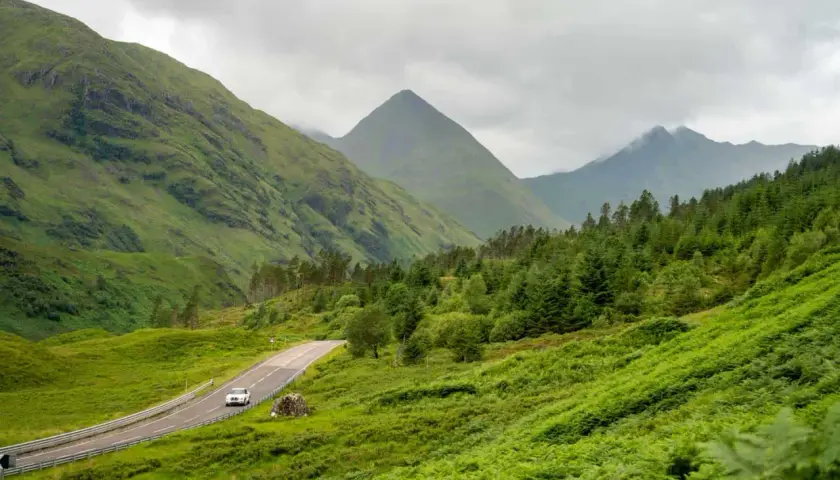 A car drives on a winding road through lush green Scottish Highlands, perfect for a hiking itinerary.