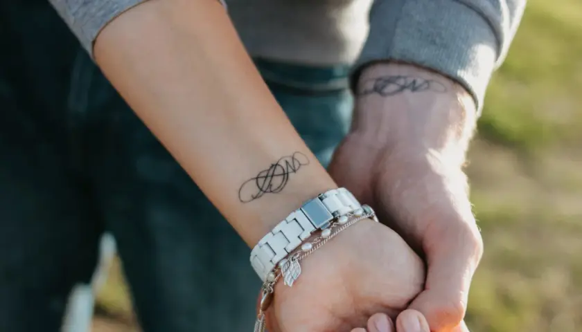 Dwa nadgarstki splecione w uścisku, każdy z subtelnym tatuażem. Na jednym widoczny jest symbol nieskończoności, na drugim ozdobny wzór.