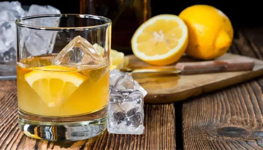 Ein erfrischender Penicillin Cocktail mit Eiswürfeln und Zitronenscheibe auf Holztisch. Daneben liegen frische Zitronen.