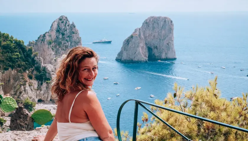 Donna sorridente ammira i Faraglioni, una delle cose da vedere a Capri, con il mare azzurro e barche.