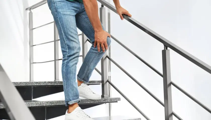 Mann mit Jeans und grünem Shirt hält sich das Knie, während er eine Treppe hinuntergeht. Er hat Probleme beim Treppen runter gehen.