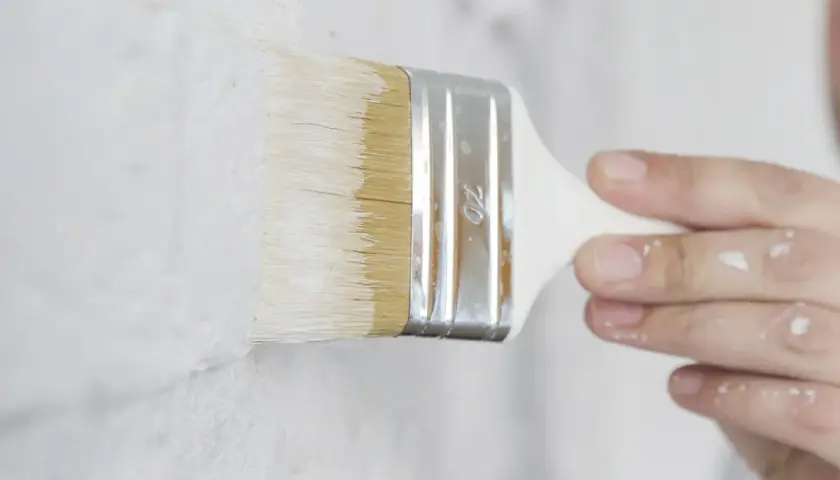 Pennello bianco stende pittura su muro grezzo. Si può mischiare il fissativo nella pittura per una finitura migliore.