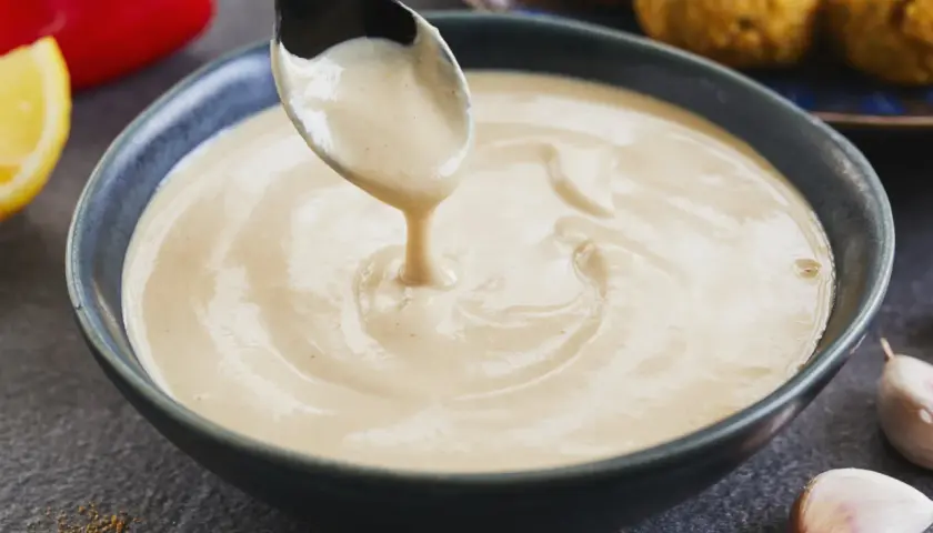 Cremige Tahini-Sauce tropft von einem Löffel in eine Schüssel. Sie ist die perfekte Begleitung zu Falafel, wie sie im Hintergrund zu sehen sind.