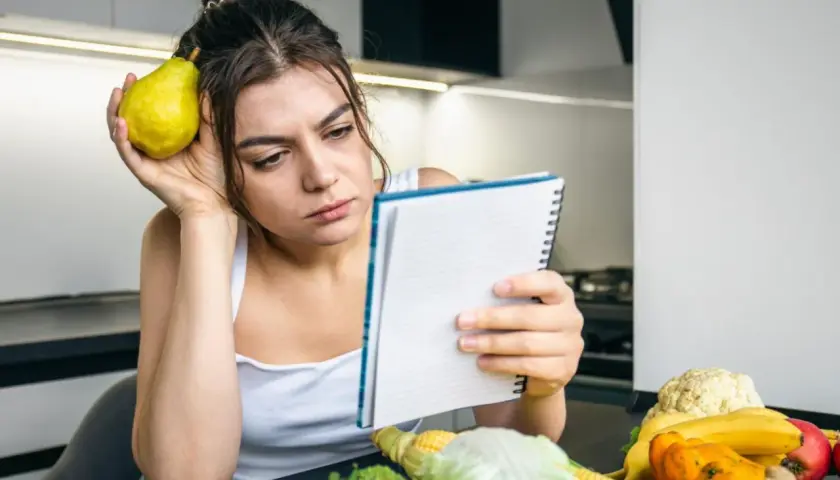 Zamyślona kobieta z gruszką przy uchu przegląda notatnik. Na stole warzywa i owoce, jakby planowała dietę 500 kcal.