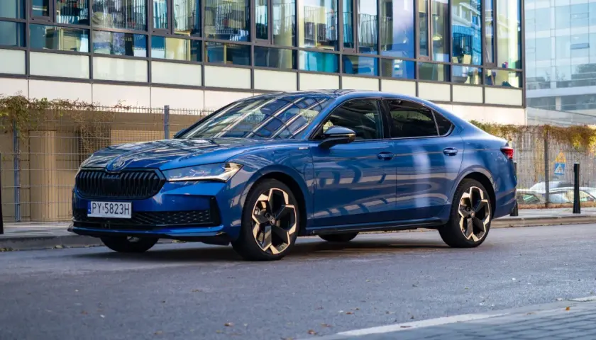 Elegancka, błękitna Skoda Superb 2025 z nowoczesnymi felgami stoi przed szklanym budynkiem.