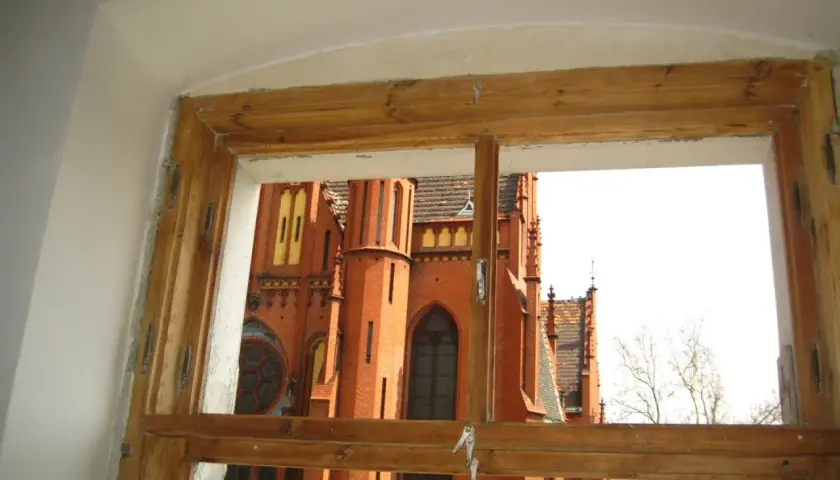 Widok przez stare okna skrzynkowe na ceglaną, zabytkową budowlę z wieżami i ozdobnymi detalami.