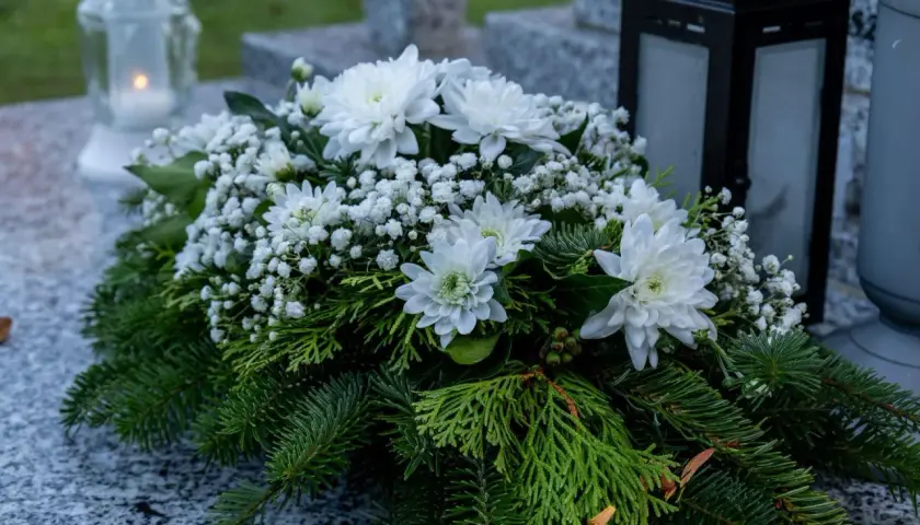 Wieniec pogrzebowy z białymi chryzantemami, gipsówką i gałązkami iglaków na kamiennym nagrobku.