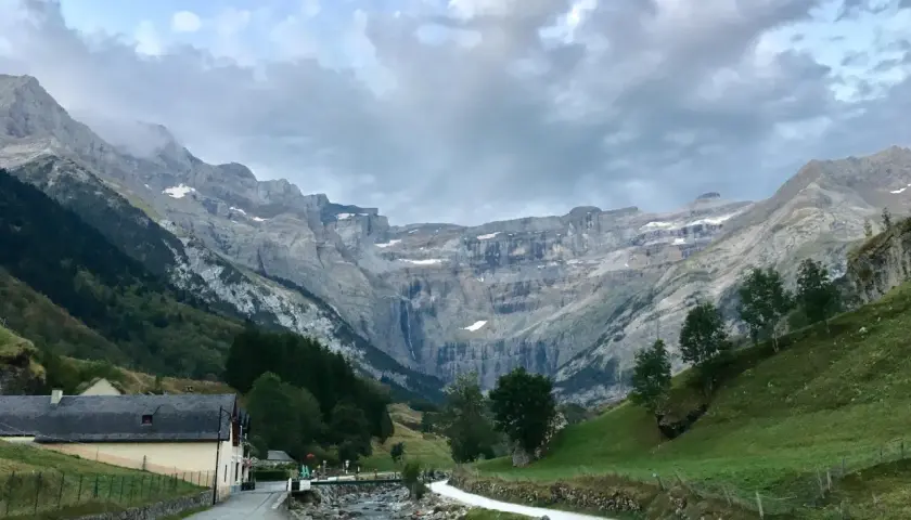 Un camino serpentea junto a un río rocoso hacia el imponente pico Taillón, con una cascada visible en la distancia y un pueblo a la izquierda.