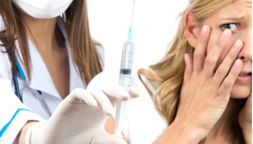 Una donna con espressione di paura degli aghi, copre il viso mentre un'infermiera con guanti bianchi tiene una siringa.