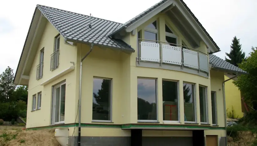 Nowy dom z żółtą elewacją i dachem w kształcie litery L. Duże okna i balkon dodają mu nowoczesnego charakteru.