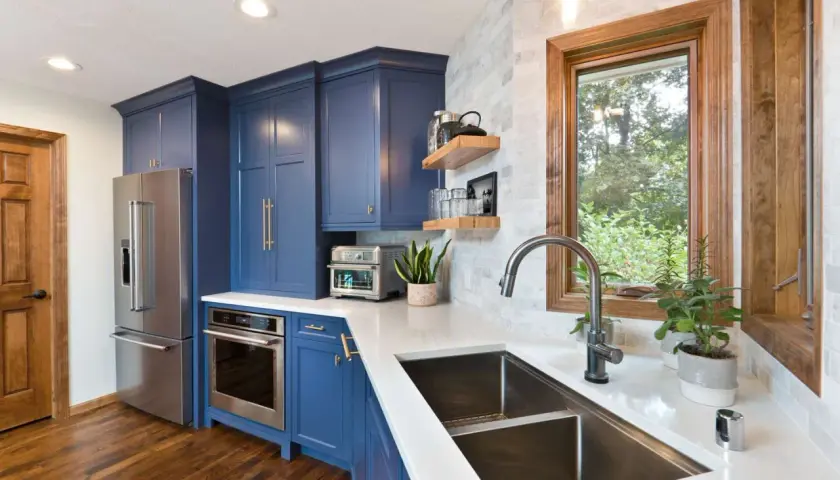 Stylish kitchen sink styling with a modern faucet, double basin sink, and a window overlooking greenery. Blue cabinets and stainless steel appliances complete the look.